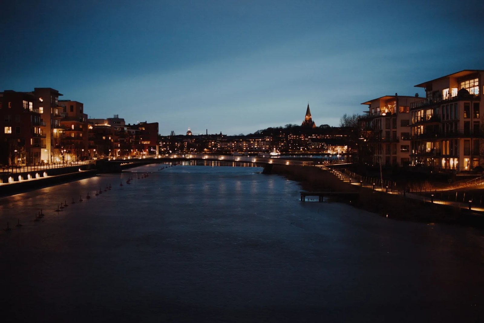 A river at dusk, lined with lit apartment buildings, a church spire on the far bank.